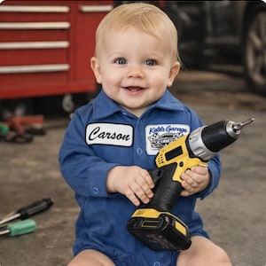 Può includere: Un bambino sorridente con una tuta blu da meccanico, che tiene in mano un trapano avvitatore giallo e nero. La tuta ha una toppa con il nome "Carson" e un'altra con "Kate's Garage".