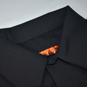 Puede incluir: Una camisa negra lisa con cuello de punta. En el interior del escote se observa una etiqueta rectangular de color naranja que muestra parcialmente el texto "TO KAP" en letras negras. La tela tiene un aspecto liso y mate.