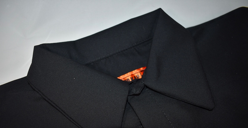 Puede incluir: Una camisa negra lisa con cuello de punta. En el interior del escote se observa una etiqueta rectangular de color naranja que muestra parcialmente el texto "TO KAP" en letras negras. La tela tiene un aspecto liso y mate.