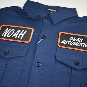 May include: A navy blue work shirt with two patches. The left patch says "NOAH" in white lettering on a black background with an orange border. The right patch says "DEAN AUTOMOTIVE" in white lettering on a black background with an orange border.