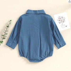 May include: A long-sleeved blue romper featuring a classic collar, a button-down front, and cuffed sleeves with single buttons. The garment is displayed from the back, showcasing its solid blue fabric and simple, structured design.