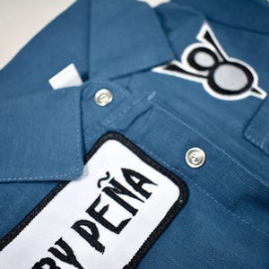 May include: A blue button-down shirt featuring a white rectangular patch with the black text "PE&Ntilde;A" and a secondary patch on the chest displaying a black and white circular emblem. The shirt includes a classic collar and silver-toned metal snap buttons.