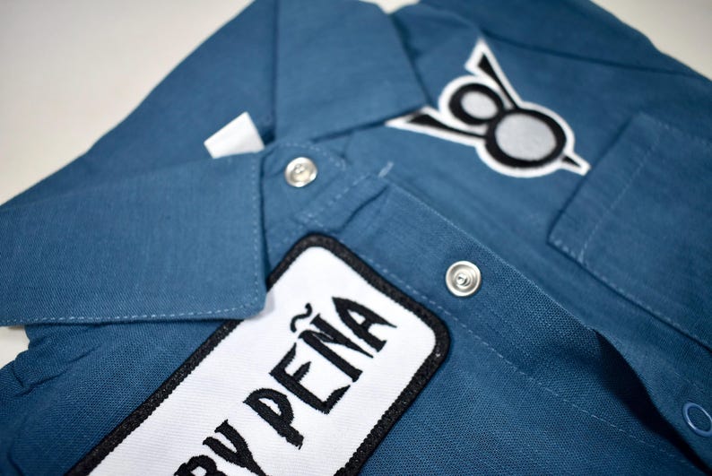 May include: A blue button-down shirt featuring a white rectangular patch with the black text "PE&Ntilde;A" and a secondary patch on the chest displaying a black and white circular emblem. The shirt includes a classic collar and silver-toned metal snap buttons.