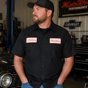May include: A black short-sleeved button-down work shirt featuring two rectangular patches on the chest. The left patch displays the name Anthony in red script, and the right patch displays the text MotoWorz in red script, both with orange borders.