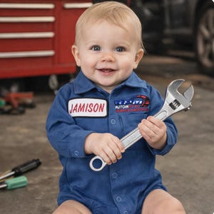 May include: A smiling baby wearing a blue mechanic's jumpsuit with the name "Jamison" on a patch. The jumpsuit has a logo that says "Automotive Service and Spares". The baby is holding a silver adjustable wrench.