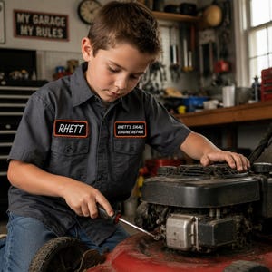 May include: A dark gray short-sleeved button-down work shirt featuring two embroidered patches. The left chest patch displays the name RHETT in white text, and the right chest patch reads RHETT'S SMALL ENGINE REPAIR in white text.