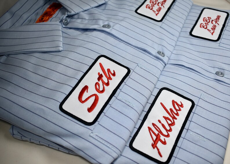 May include: A blue striped shirt with three white rectangular patches. The patches have black borders and red cursive text. The text on the patches reads "Seth", "Alisha", and "Bobo Law Firm".