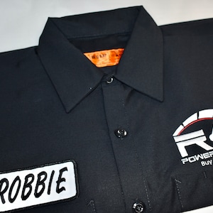 Peut inclure: Chemise de travail noire avec une étiquette de nom blanche qui dit "ROBBIE" et un logo rouge, blanc et noir qui dit "RG POWERSPORTS+ BUY/SELL/TRADE"