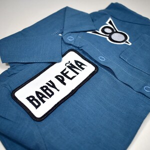 May include: A blue collared shirt featuring a rectangular white patch with the black text BABY PE&Ntilde;A and a smaller embroidered patch above the chest pocket displaying a stylized black and white graphic design with two circular elements.