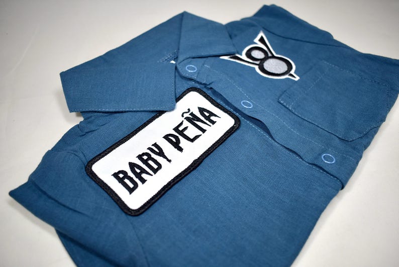 May include: A blue collared shirt featuring a rectangular white patch with the black text BABY PE&Ntilde;A and a smaller embroidered patch above the chest pocket displaying a stylized black and white graphic design with two circular elements.