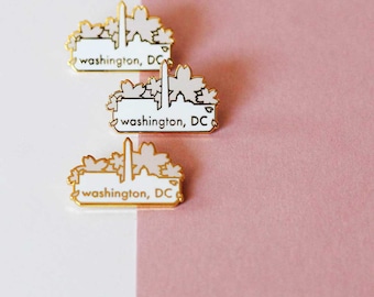 May include: Three gold-colored enamel pins with a white background. Each pin features a stylized image of the Washington Monument and cherry blossoms with the text "washington, DC".
