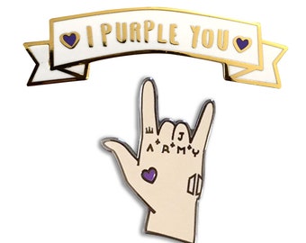 May include: Two enamel pins featuring the phrase "I Purple You" with a purple heart on a white banner and a hand sign with the word "ARMY" and a purple heart.