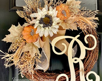 May include: A fall-themed wreath with a burlap bow, faux sunflowers, and small orange pumpkins. The wreath is made of grapevine and has a large wooden letter "R" in the center. The wreath is decorated with dried leaves and twigs.