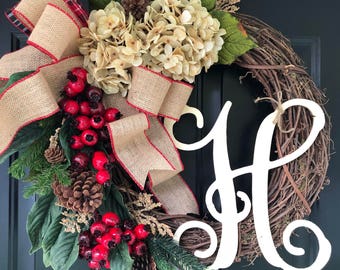 May include: A brown twig wreath with a burlap bow and red berries. The wreath is decorated with white hydrangeas, pine sprigs, and pine cones. A white wooden letter "H" is attached to the wreath.