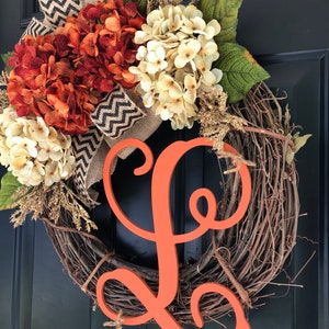 May include: A grapevine wreath decorated with orange and white hydrangeas, burlap ribbon with a black and white chevron pattern, and a large orange wooden letter L.