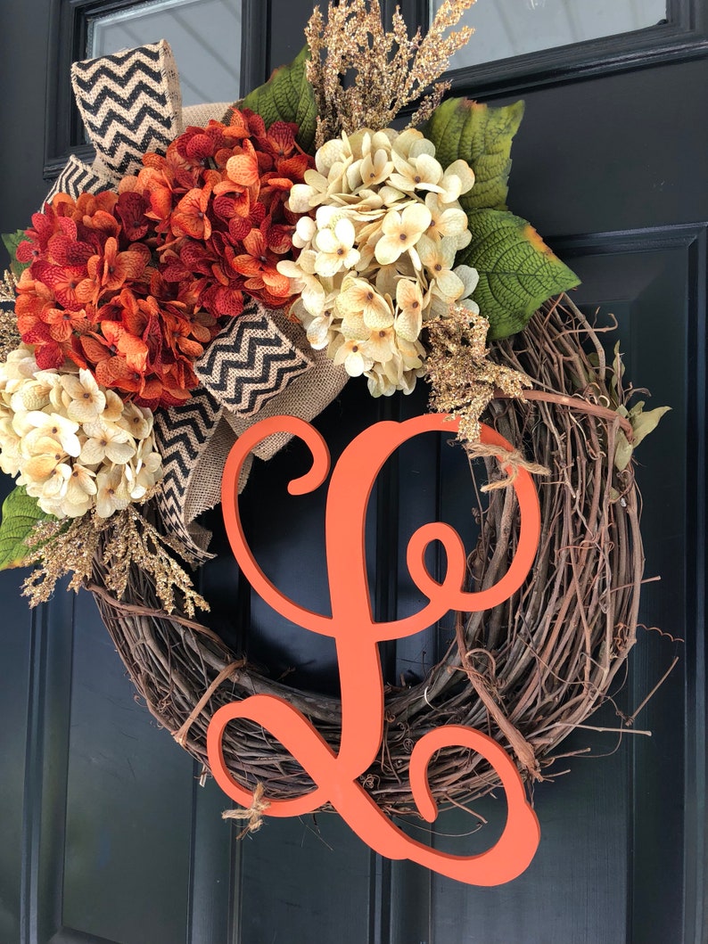 May include: A grapevine wreath decorated with orange and white hydrangeas, burlap ribbon with a black and white chevron pattern, and a large orange wooden letter L.