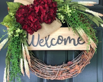 May include: A welcome wreath with a wooden sign that says "Welcome" in black script. The wreath is made of grapevine and is decorated with burgundy hydrangeas, burlap ribbon, and green and white foliage.