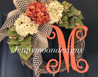 May include: A wreath with a burlap bow with black and white chevron pattern, orange and white hydrangea flowers, green leaves, and a large orange wooden letter M. The wreath is made of grapevine and has a brown twine hanging from the bottom. The text "Jennymoondesigns" is visible at the bottom of the wreath.