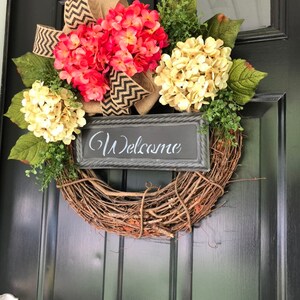 May include: A wreath made of brown twigs with a burlap bow and a sign that says "Welcome". The wreath is decorated with pink and cream hydrangeas.