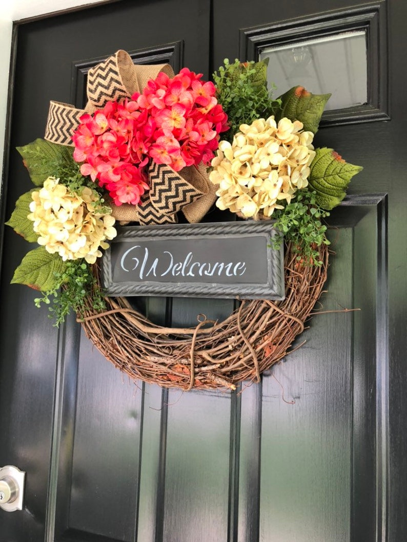 May include: A wreath made of brown twigs with a burlap bow and a sign that says "Welcome". The wreath is decorated with pink and cream hydrangeas.