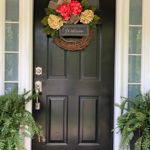 May include: A black front door with a wreath made of grapevine, pink and cream hydrangeas, and a burlap bow. The wreath has a sign that says "Welcome".
