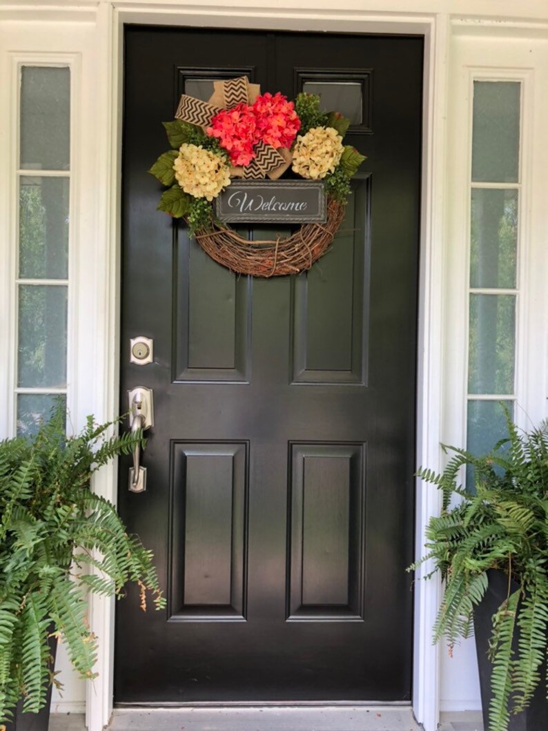 May include: A black front door with a wreath made of grapevine, pink and cream hydrangeas, and a burlap bow. The wreath has a sign that says "Welcome".