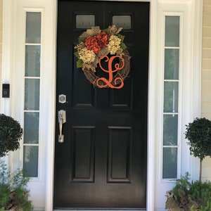 May include: A black front door with a white frame and a wreath with orange and white flowers and a large orange letter L. The door has a silver door handle and a silver door knocker. There are two potted plants on either side of the door.