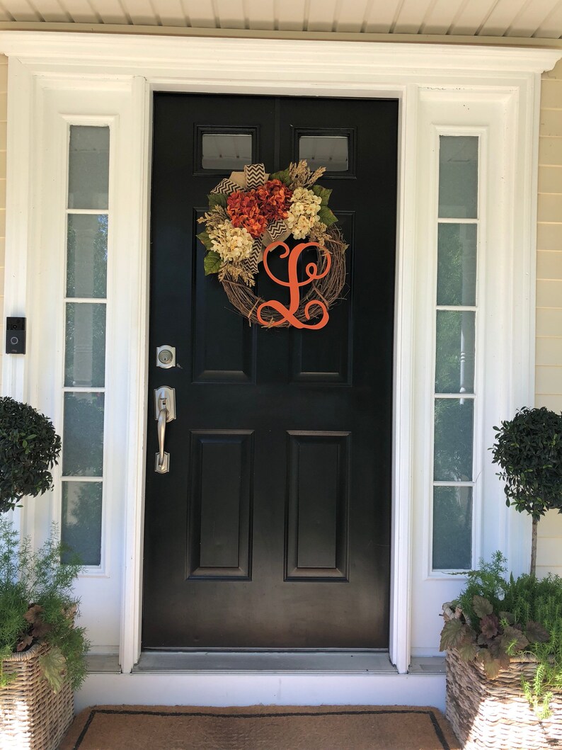 May include: A black front door with a white frame and a wreath with orange and white flowers and a large orange letter L. The door has a silver door handle and a silver door knocker. There are two potted plants on either side of the door.