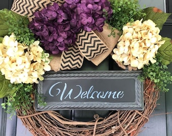 May include: A grapevine wreath with a burlap bow, purple and cream hydrangeas, and a sign that says "Welcome".
