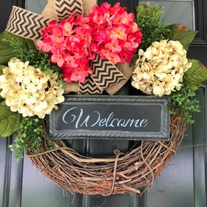 May include: A brown twig wreath with pink and cream hydrangeas, a burlap bow with black and white chevron ribbon, and a black chalkboard sign that says "Welcome".