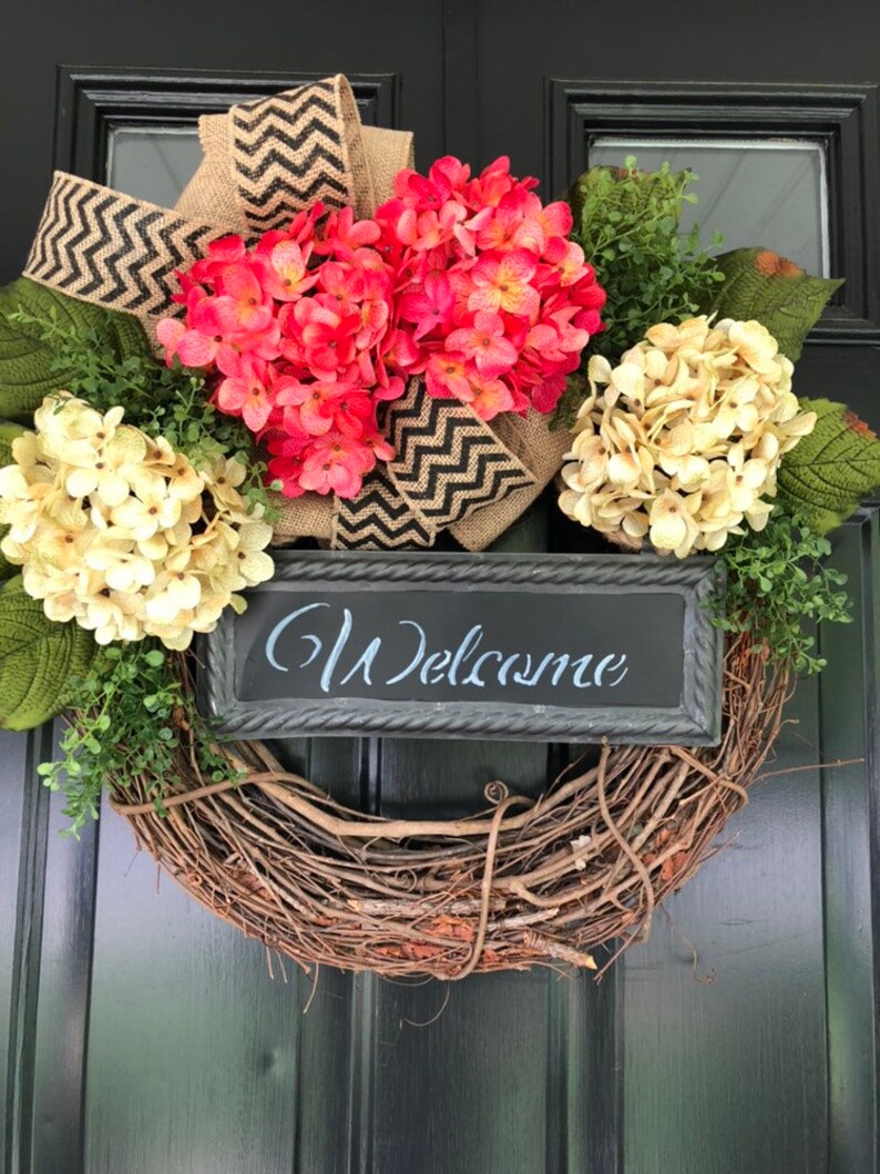 May include: A brown twig wreath with pink and cream hydrangeas, a burlap bow with black and white chevron ribbon, and a black chalkboard sign that says "Welcome".