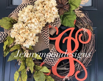 May include: A wreath made of twigs with a large orange letter "G" in the center. The wreath is decorated with burlap ribbon with black and white chevron pattern, white hydrangeas, green leaves, and a brown pinecone.