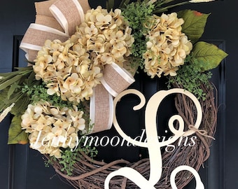 May include: A wreath made of grapevine with a large, wooden, white letter L in the center. The wreath is decorated with cream-colored hydrangeas, greenery, and a burlap bow with a white ribbon.