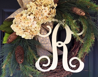 May include: A wreath with a white wooden letter "R" in the center. The wreath is decorated with pine branches, brown pine cones, and a large beige hydrangea with a brown and gold polka dot bow.