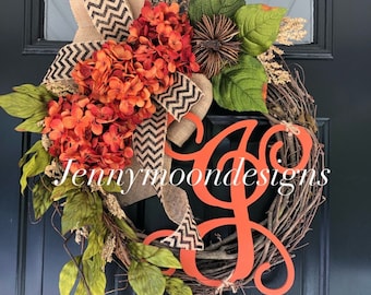 May include: A fall-themed wreath with orange hydrangeas, burlap ribbon with black and white chevron pattern, and a large orange cursive letter "J" on a twig base.