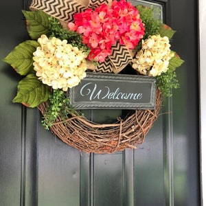 May include: A wreath made of grapevine with a burlap and black chevron ribbon bow. The wreath is decorated with pink and white hydrangeas and a black sign that says "Welcome".