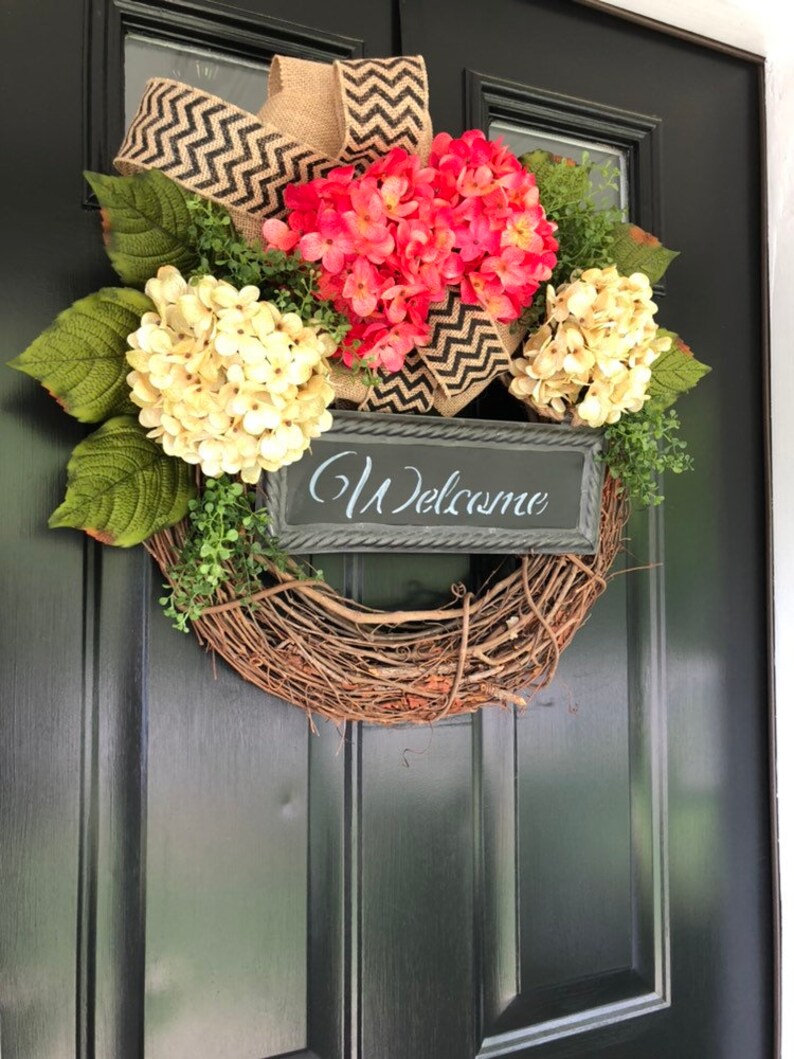 May include: A wreath made of grapevine with a burlap and black chevron ribbon bow. The wreath is decorated with pink and white hydrangeas and a black sign that says "Welcome".