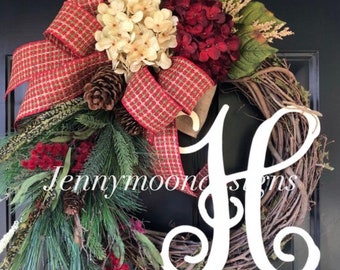 May include: A Christmas wreath with a red and green plaid bow, pine branches, red berries, and a white wooden letter H. The wreath is made of grapevine and has a rustic look.