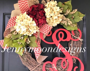 May include: A red and white chevron ribbon, burlap, and greenery surround a wreath with white and red hydrangeas. A large red wooden letter "S" is attached to the wreath. The wreath is on a black door with the text "Jennymoondesigns" visible.