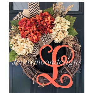 May include: A wreath with a large orange letter L in the center. The wreath is made of grapevine and is decorated with orange and white hydrangeas, burlap, and a black and white chevron ribbon. The wreath is hanging on a black door. The text "Jennymoondesigns" is visible at the bottom of the image.
