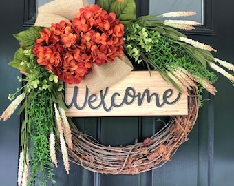 May include: A welcome wreath with a wooden sign that says "Welcome" in black script. The wreath is made of grapevine and is decorated with orange hydrangeas, burlap, and green and white foliage.
