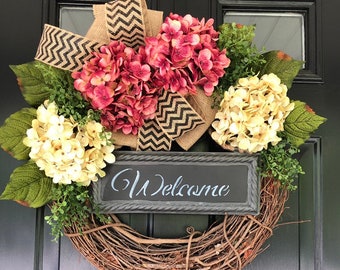 May include: A grapevine wreath with pink and cream hydrangeas, a burlap bow with black and white chevron print, and a black chalkboard sign that says "Welcome".