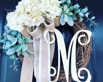 May include: A white and green wreath with a large white letter "M" in the center. The wreath is decorated with white hydrangeas and green leaves. The letter "M" is made of wood and has a rustic finish.