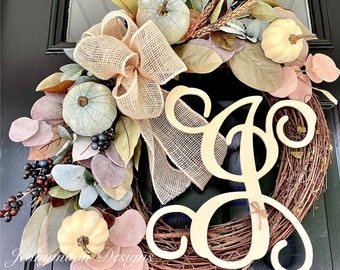 May include: A fall-themed wreath with a burlap bow, faux greenery, and white pumpkins. The wreath is decorated with a wooden sign that says "&" in a cursive font.