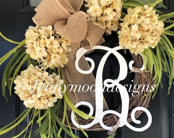 May include: A grapevine wreath adorned with cream-colored hydrangea blooms, green foliage, and a burlap bow. A large white letter "B" is centered on the wreath. The text "jennymoodesigns" is visible on the bottom left.
