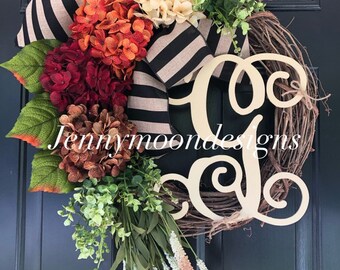 May include: A wreath with a wooden "C" monogram, decorated with black and white striped ribbon, burgundy, orange, and cream hydrangeas, greenery, and pink sprigs.