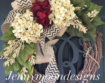 May include: A wreath made of grapevine with a burlap bow and a black monogram "R". The wreath is decorated with white hydrangeas, red flowers, and green leaves.