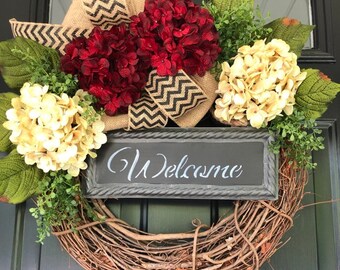 May include: A grapevine wreath with a burlap bow, red and cream hydrangeas, and a black and white chevron ribbon. A black chalkboard sign with the word "Welcome" is attached to the wreath.