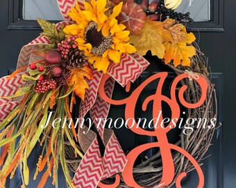 May include: A fall-themed wreath with a large orange monogram "G" in the center. The wreath is decorated with sunflowers, fall leaves, berries, and a red and brown chevron ribbon. The wreath is on a black door with the text "Jennymoondesigns" visible.