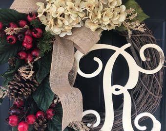 May include: A brown burlap wreath with a white hydrangea, red berries, pine cones, and a white wooden letter "R" monogram.
